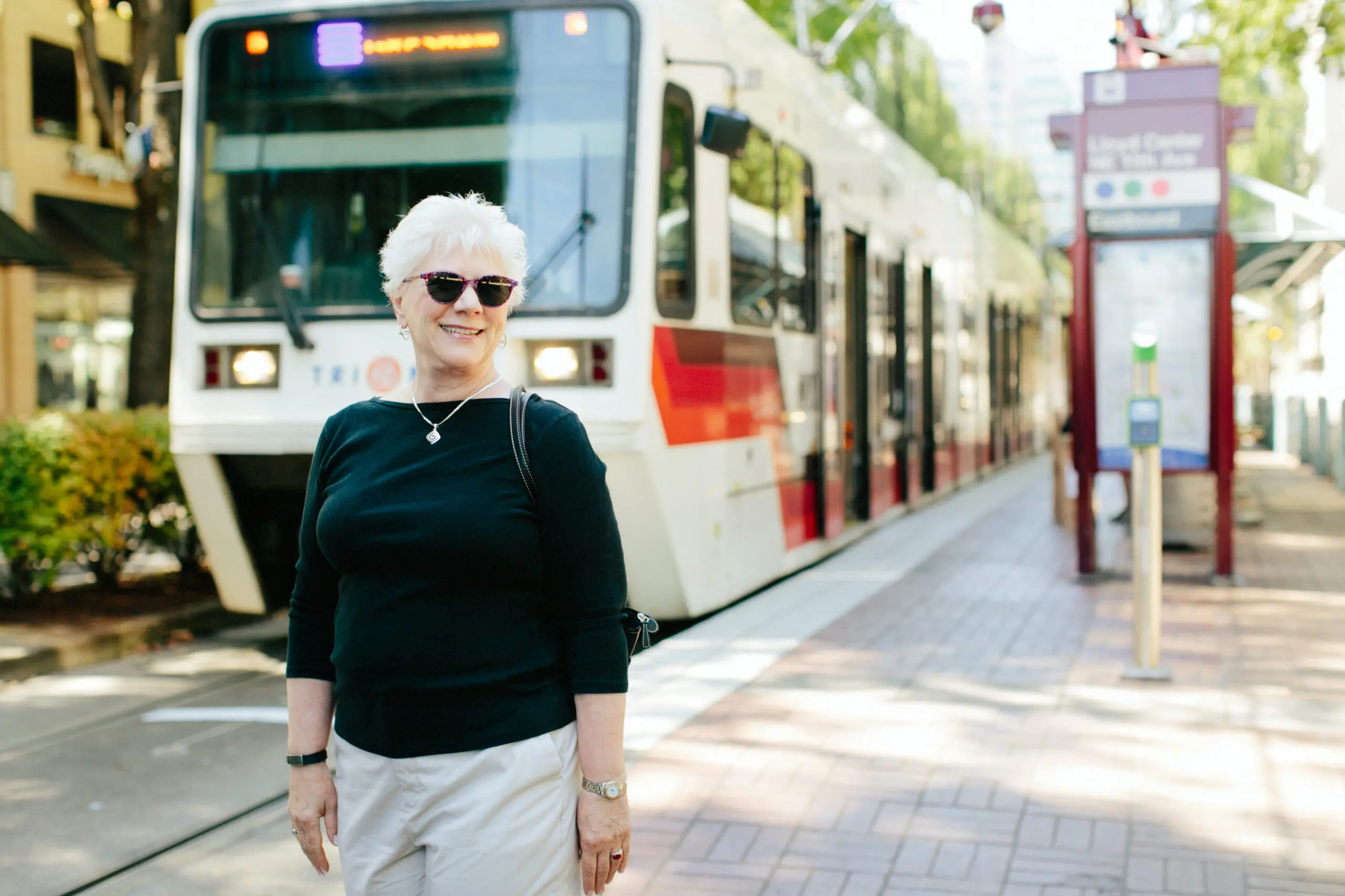 A smiling older woman wearing sunglasses stands in front of a streetcar.